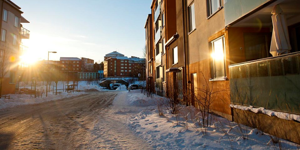 En gata med flerbostadshus på vardera sidan och med en pendeltågsstation i bakgrunden. Det är vinter och gatan är snöröjd. Solen ligger lågt och skiner mellan husen. Foto.