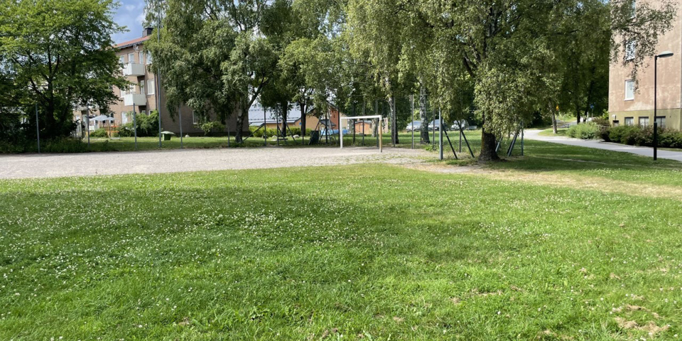 En grusbollplan med fotbollsplan intill en gräsmatta. Bolplanen är delvis inhägnad och bakom planen ses höga träd och flerbostadshus. Foto
