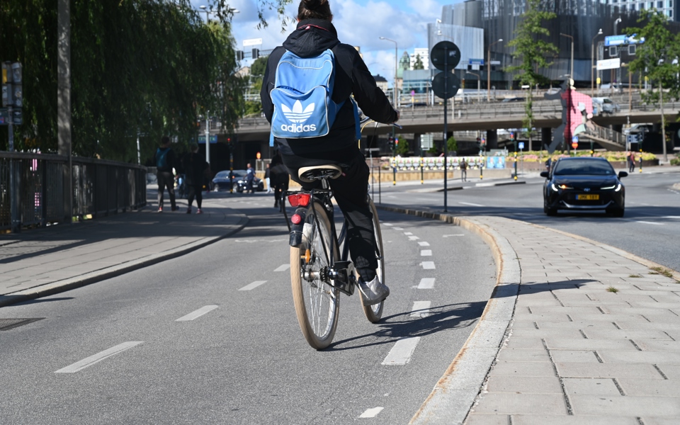 Cyklande person med ryggsäck sedd bakifrån på en cykelbana. I bakgrunden syns en bil, byggnader och människor i rörelse, fotografi.