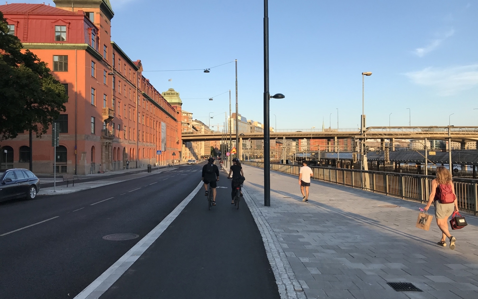 Cykelbana, bilväg och trottoar, vy mot bro i bakgrunden. Byggnad, tågspår och människor i rörelse, fotografi.
