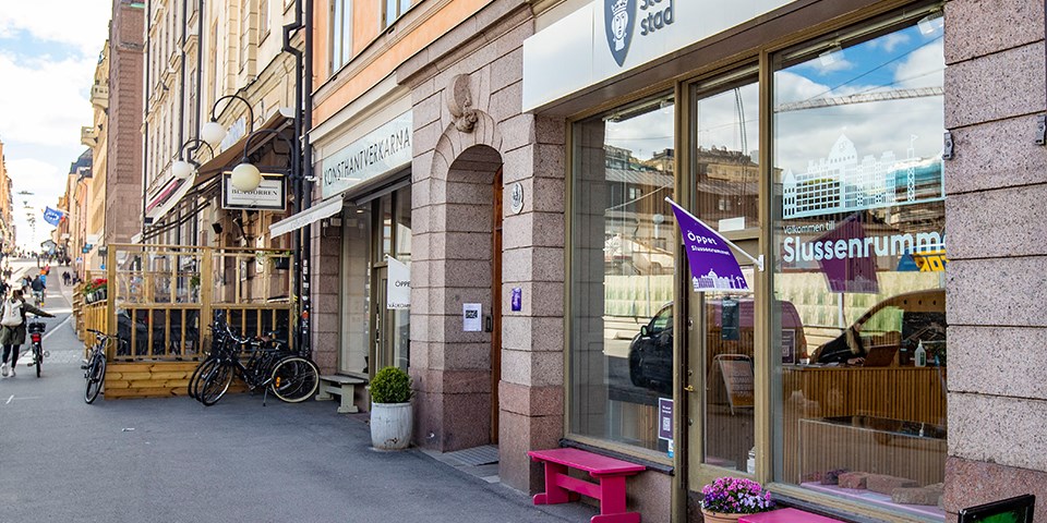 Fasad utanför utställningslokalen Slussenrummet. Skyltfönster och entré med rosa bänkar och en blomkruka med rosa blommor utanför. Vid entrén hänger en lila flagga med texten "Öppet". Ovanför entrén sitter en skylt med Stockholms stads logo.