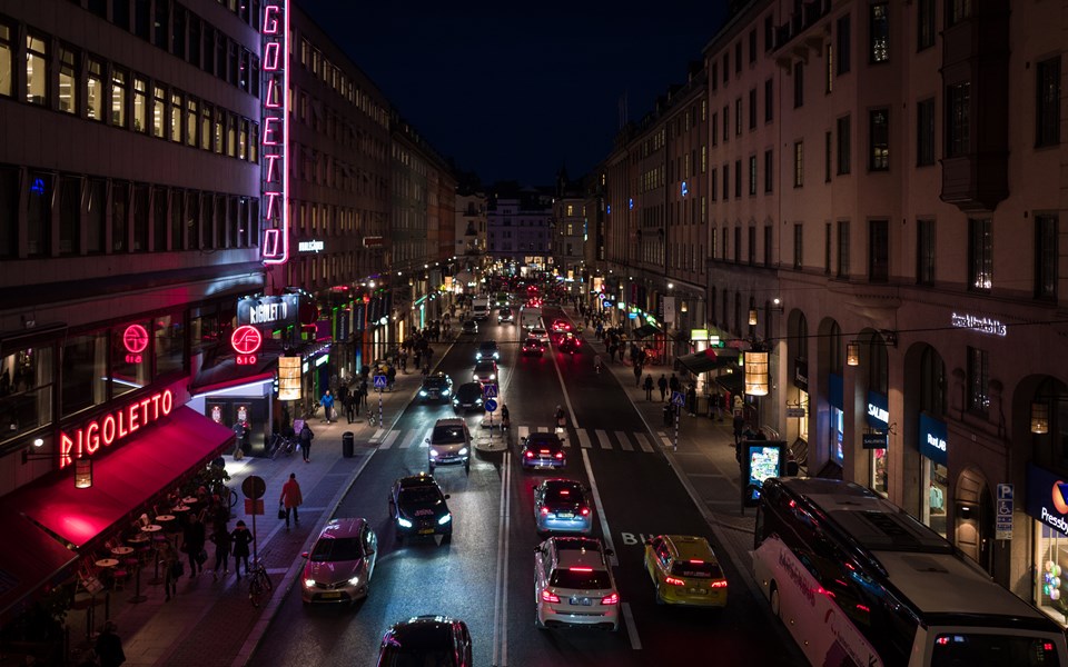 Kungsgatan kvällstid. Männiksor går och bilar kör. Neonskyltar lyser på husen.